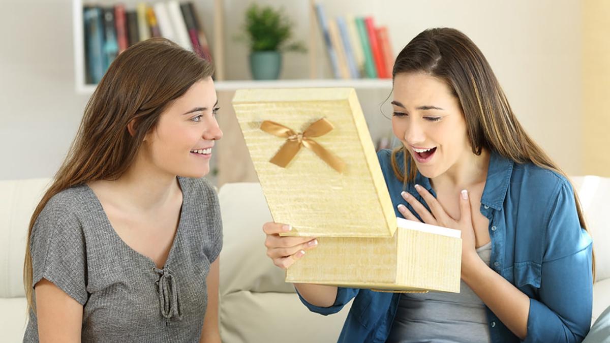 Article Cards Featured Image Two happy friends opening a gift box sitting on a couch in the living room at home