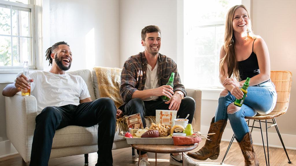 A photo of the big game with three people sitting in a living room watching a tv and laughing while holding beers with a plate of snacks in front of them.