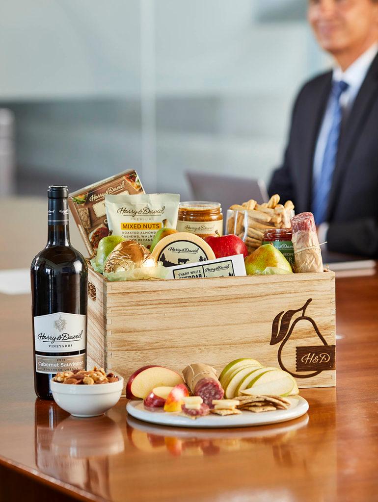 A photo of employee appreciation day with a gift box full of food and snacks on a conference table next to a bottle of wine with a man in a suit in the background.