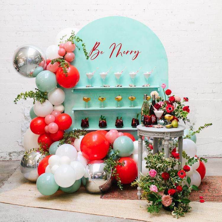 Holiday cocktail wall and drink station full of multiple glasses of different varieties of cocktails surrounded by holiday colored balloons and flowers.